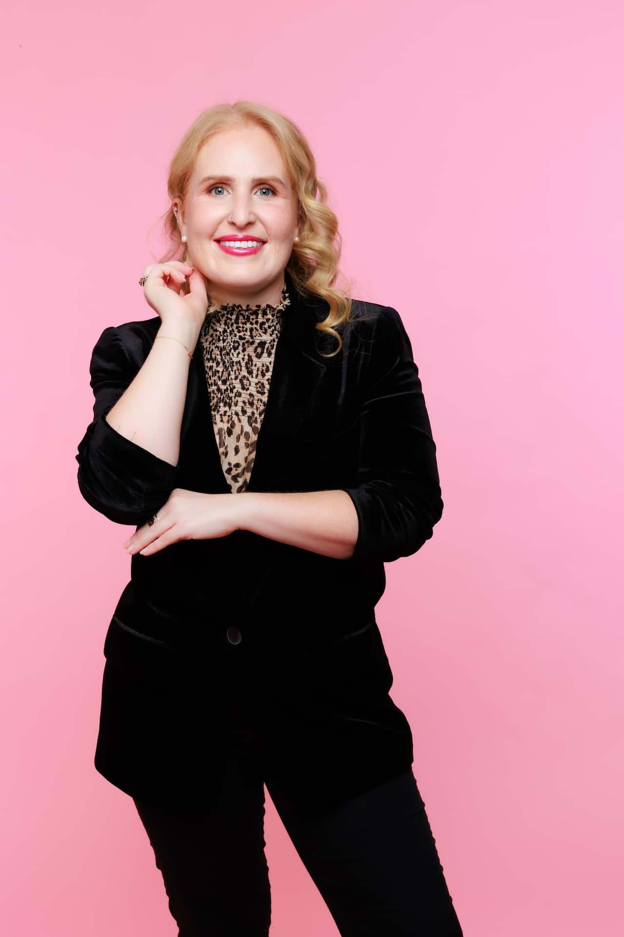 A woman in a black suit smiles at the camera against a pink background.