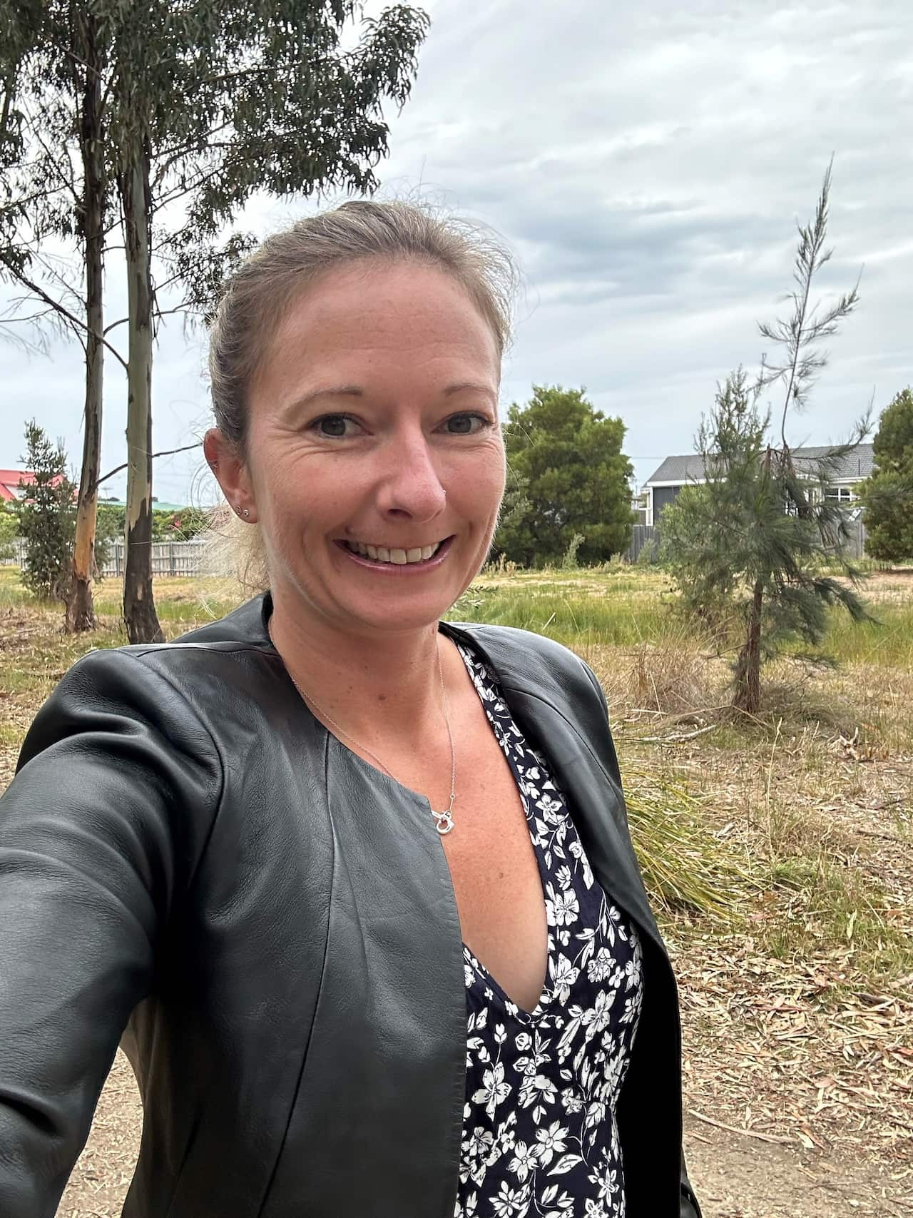 a woman smiling wearing a black jacket takes a selfie while standing outside 