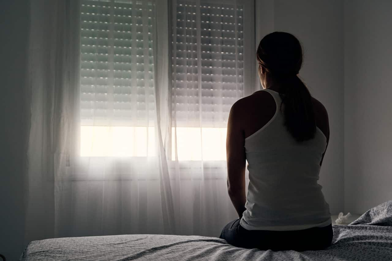 Rear view of a woman sitting on her bed looking out the window.