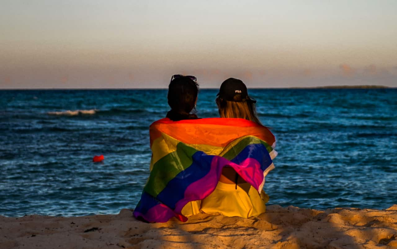 Two people hugging, wrapped in a rainbow flag on a cliffside looking over an ocean 