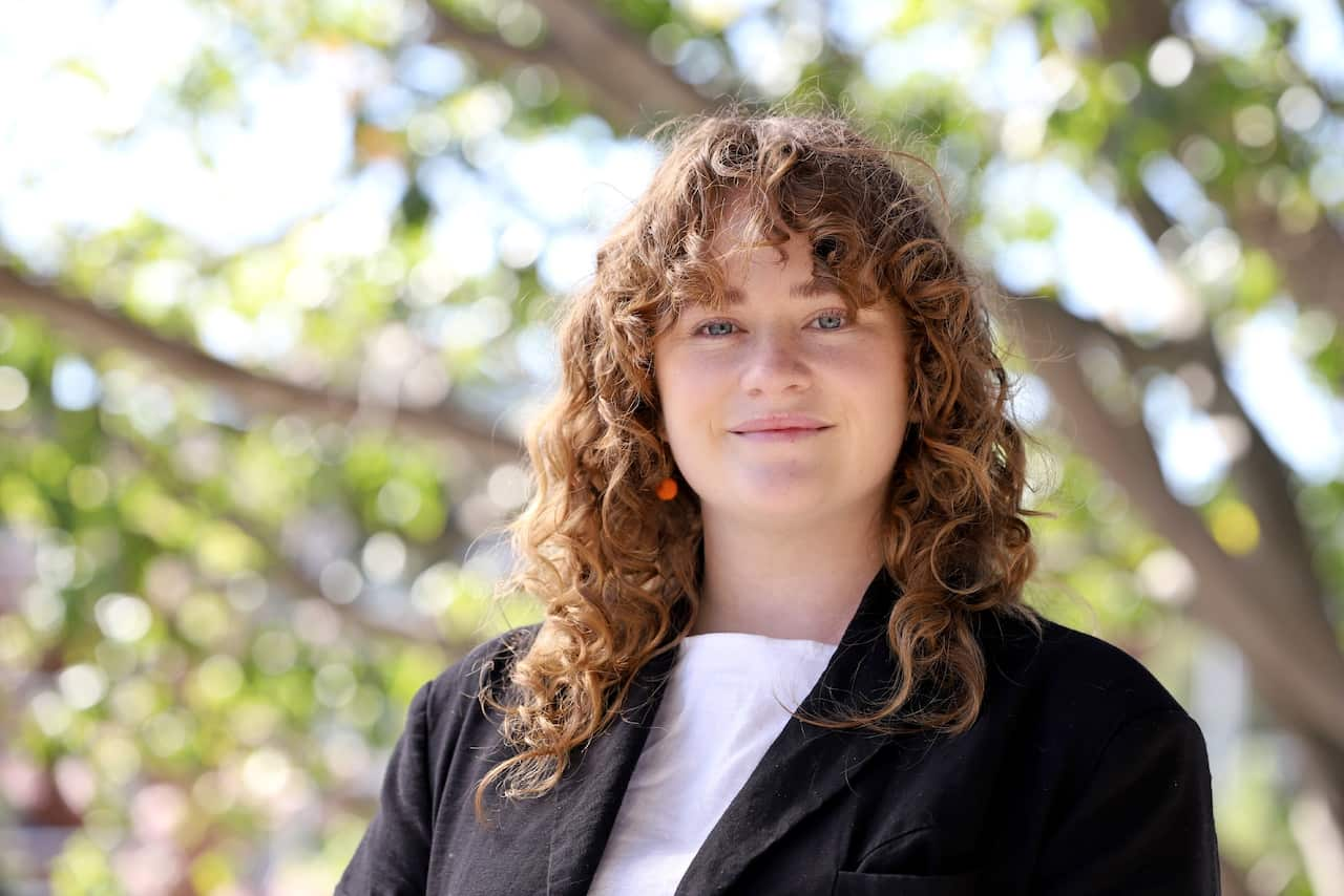 A woman with long curly brown hair wearing a dark jacket and white t-shirt standing outside smiling