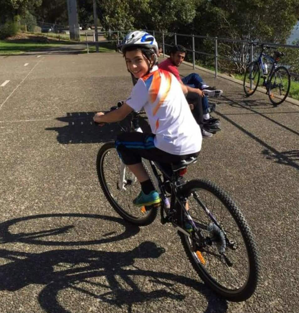 A boy in his early teens wearing a white t-shirt and a bike helmet riding a bike and looking over his left shoulder at the camera