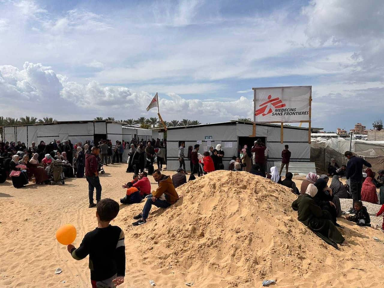 Adults and children sitting on a sand pile outside a Medecins sans Frontieres clinic.