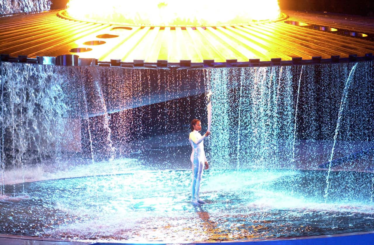 Cathy Freeman wearing a white bodysuit standing under cascading water while holding the Olympic torch.