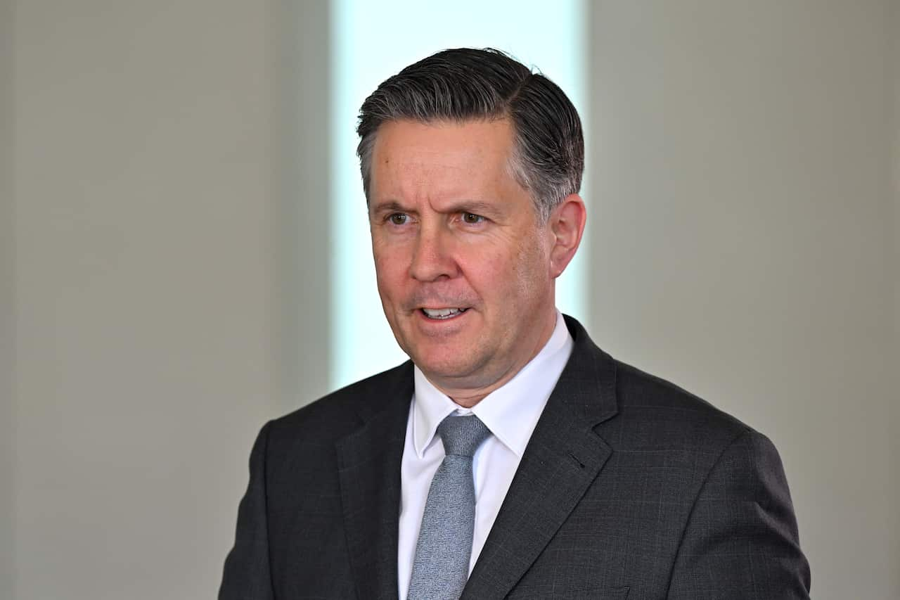 A man in a dark grey suit, a white shirt and blue tie, speaking in front of a white wall.