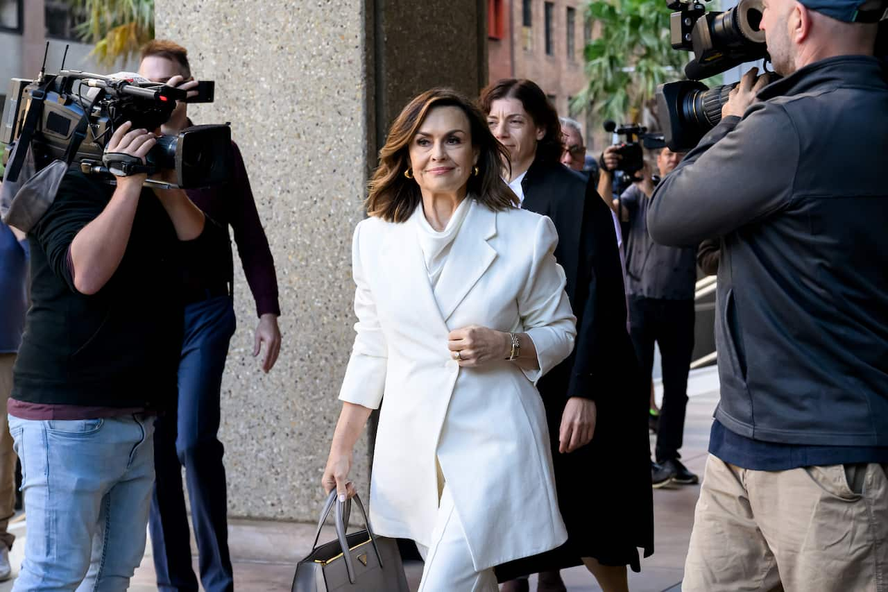 A woman in a white coat walking while surrounded by camera operators.