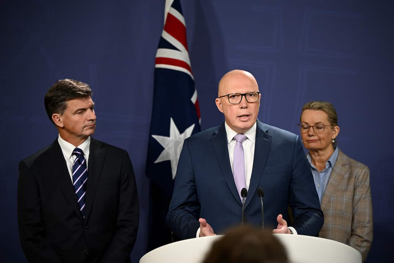 Three people standing, one is behind a podium and speaking into a microphone. An Australian flag is behind them.