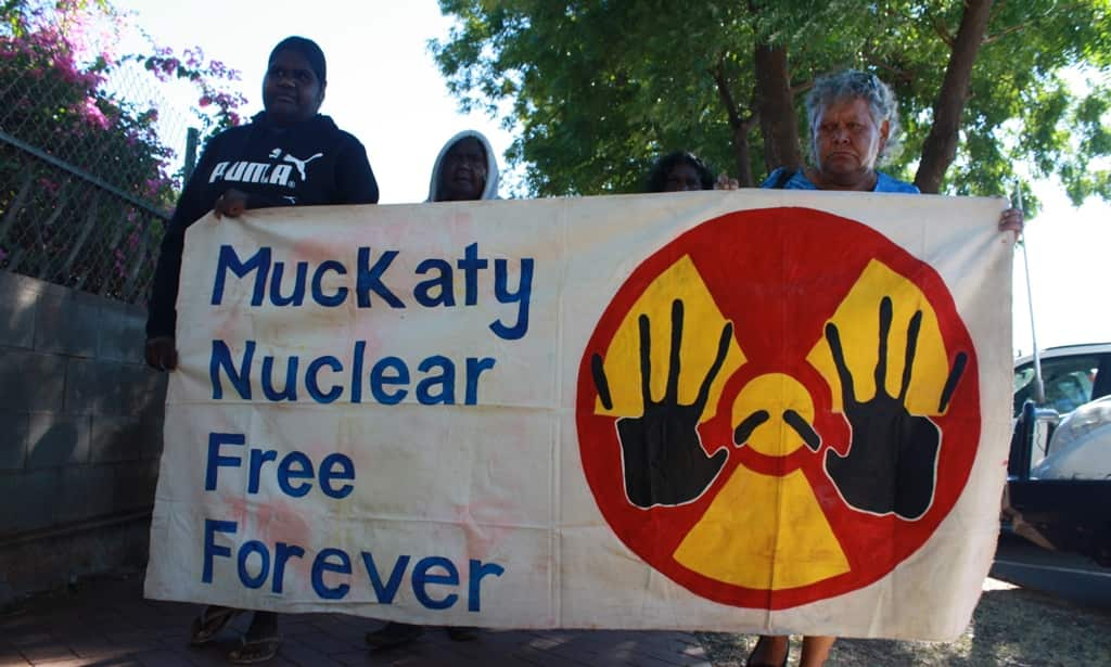 Protesters holding a banner that reads "Muckaty Nuclear Free Forever". The banner has a radioactivity symbol with two black hands superimposed over it. 