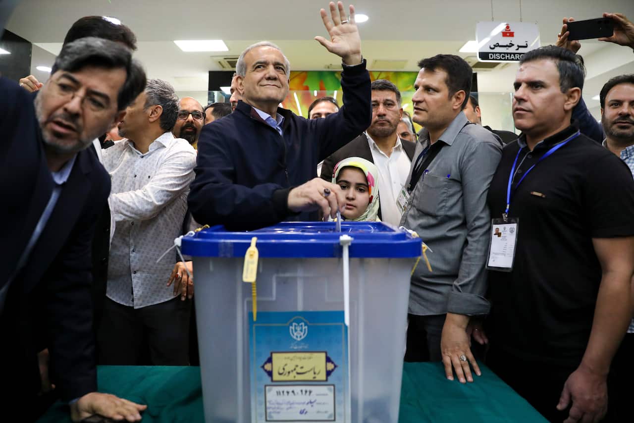 A man waving while putting a piece of paper into a ballot box. He is surrounded by other people.