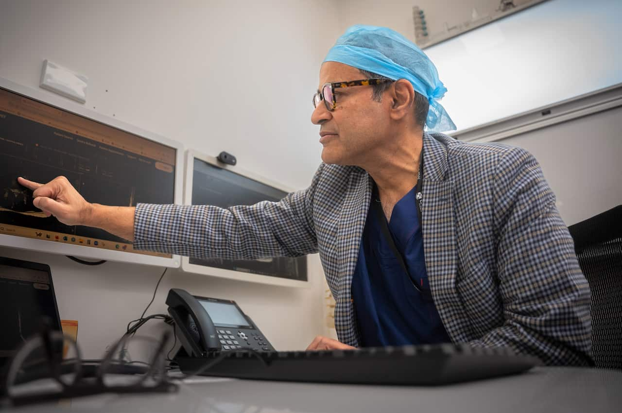 A man in a suit jacket and surgical headwear points to an image on a computer screen.