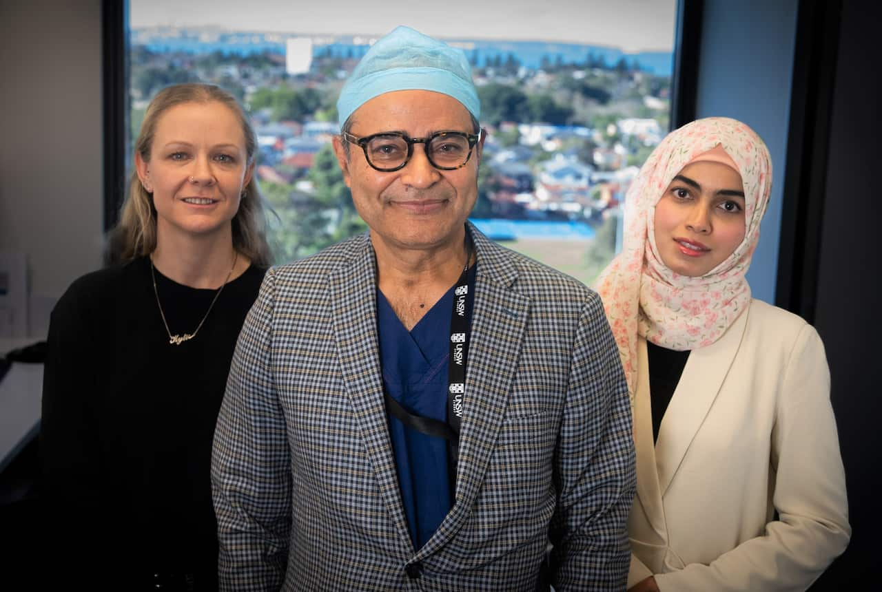 A woman in a black sweater stands next to a man in a grey suit jacket, and another woman in a head scarf. 