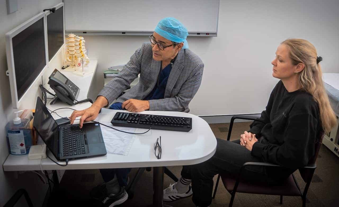 A man in  checked jacket points to a computer screen while a woman with blonde hair watches on. 