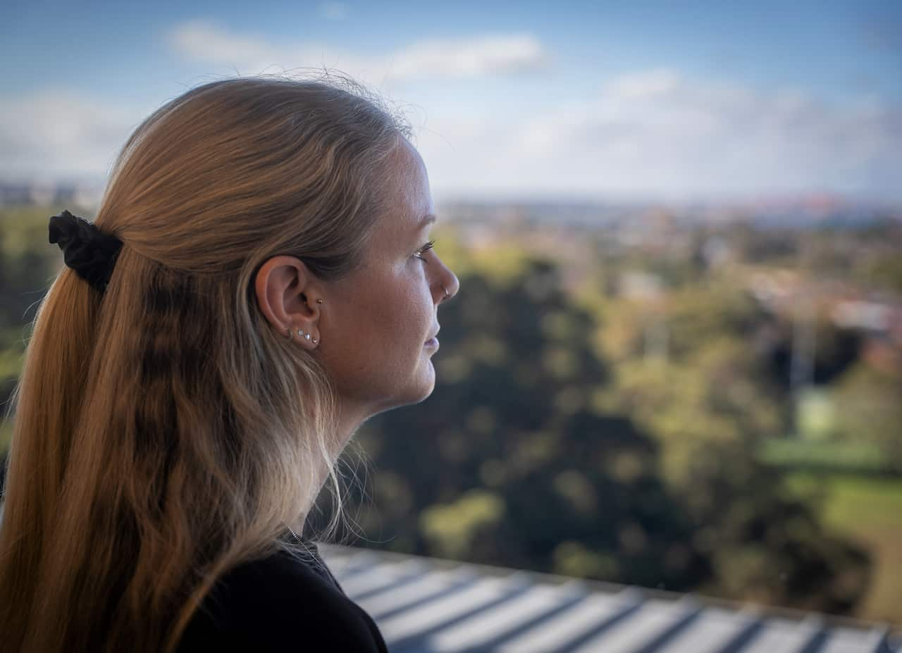 A woman with long hair stands near a window looking out at the view. 