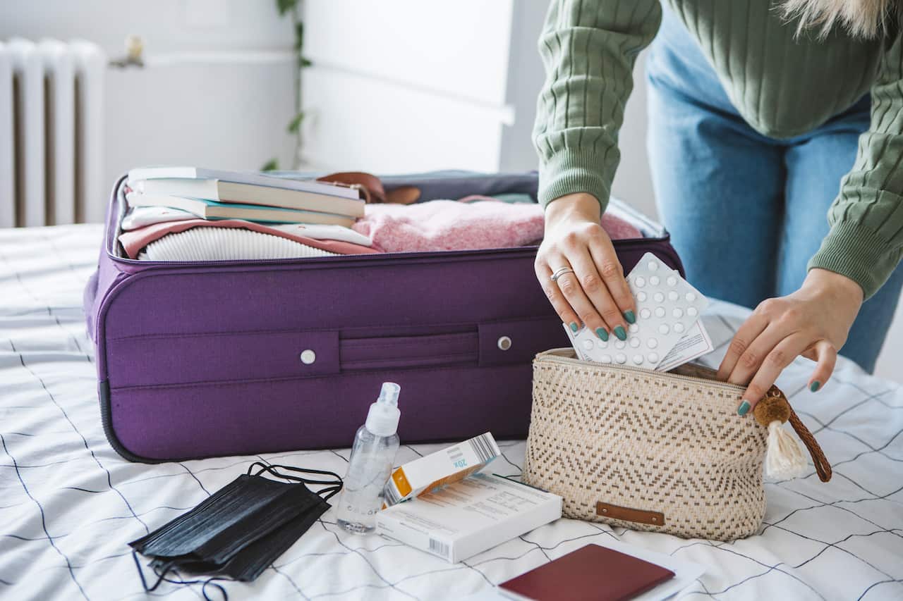 A woman packing medication in a cosmetic bag. Alongside it on the bed is an open purple suitcase, passport, black face masks, and disinfectant spray