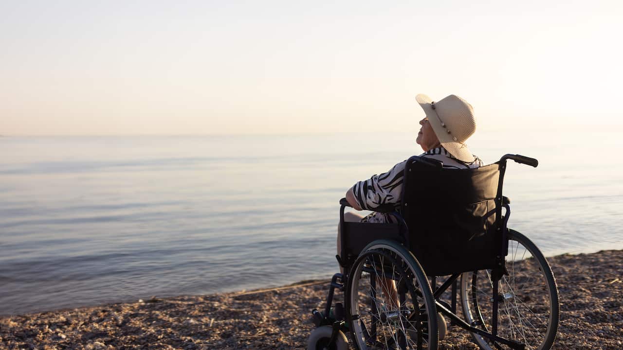 An elderly woman wearing a straw hat, sitting in a wheelchair near a lake