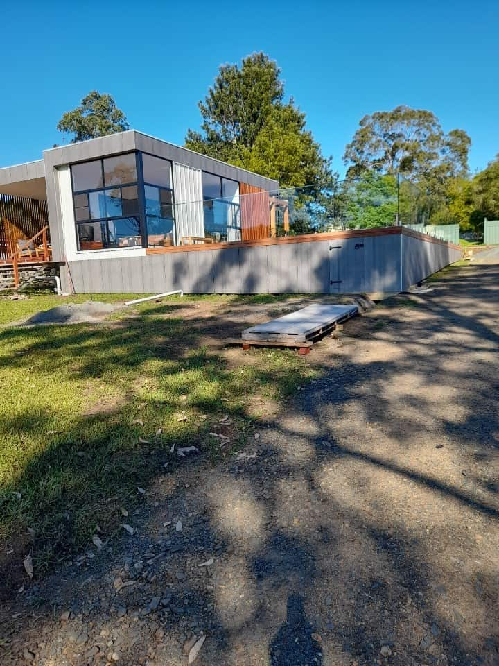 A square house with glass fencing around a deck, and some construction materials on the ground nearby