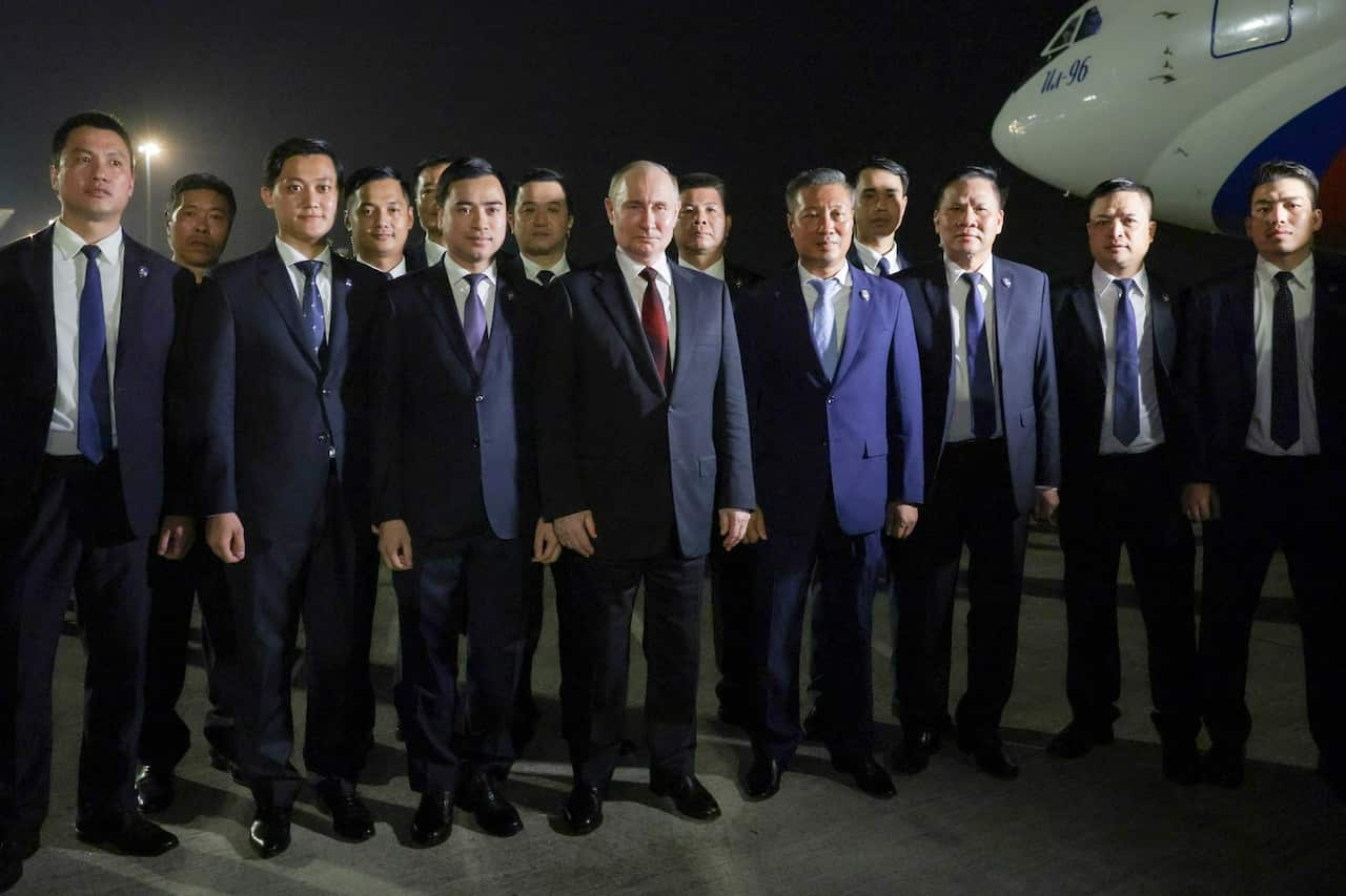 A large group of men in suits, with Vladimir Putin in the centre, standing for a photograph. An aircraft is behind them.