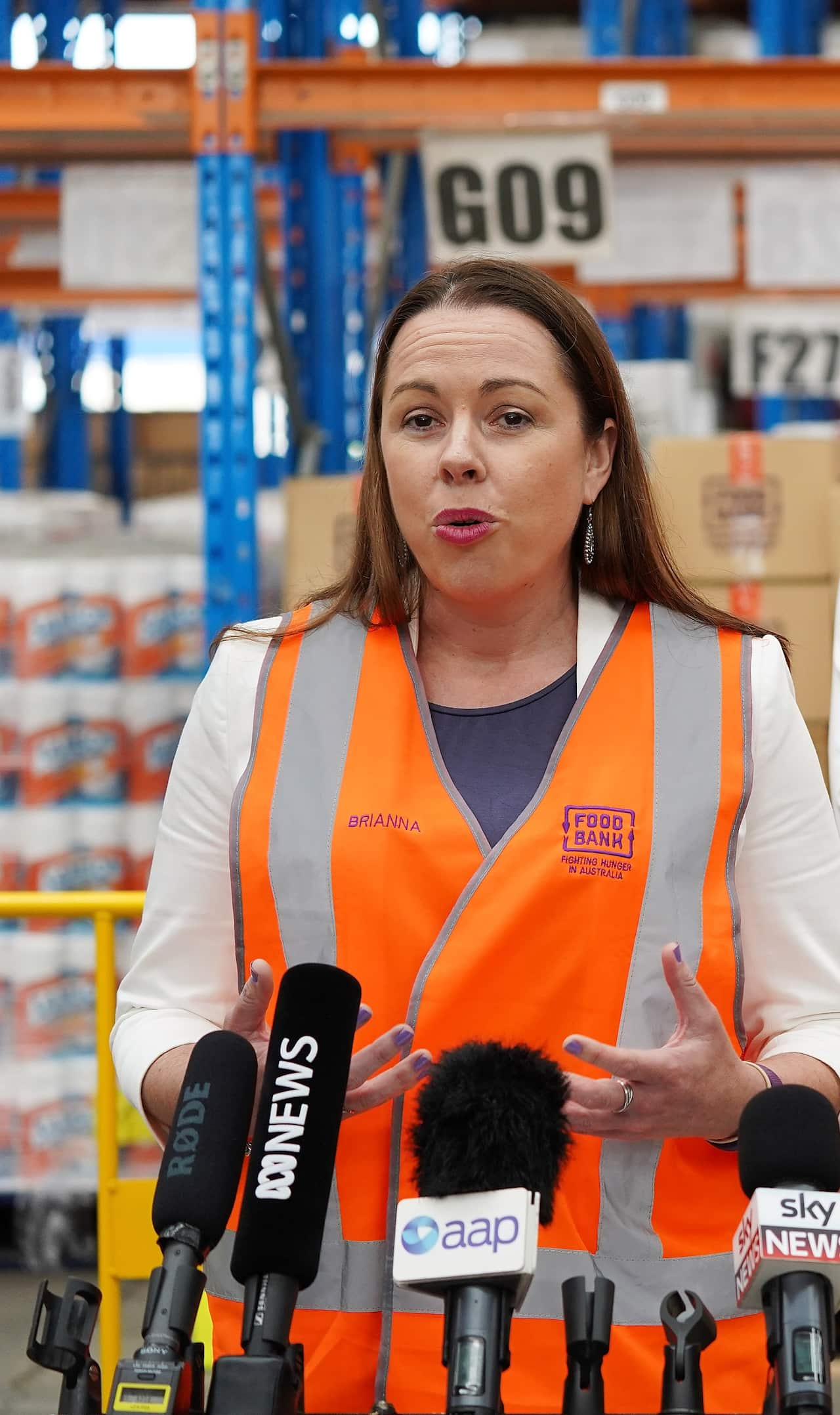 A woman wearing an orange hi-vis vest stands in a warehouse talking behind a row of microphones.