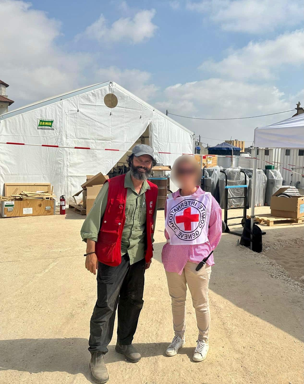 Two aid workers, one of which is wearing a Red Cross vest, whose face is blurred. They're standing on sand and there are large camp tents behind them.