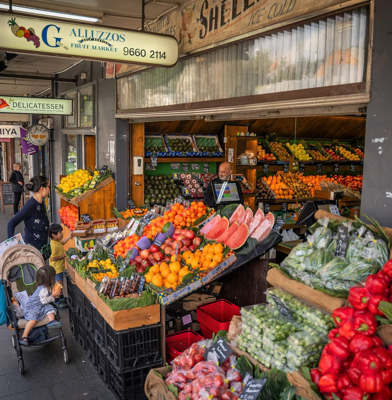 Rows of fresh fruit ad vegetables on the street as a woman with children and a stroller looks on.