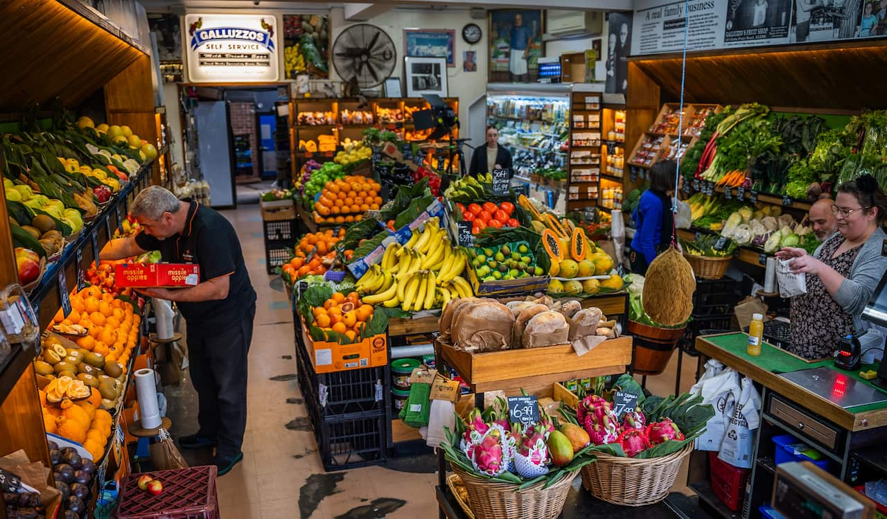 A man stocks shelves inside a fruit shop while a customer stands at the counter.