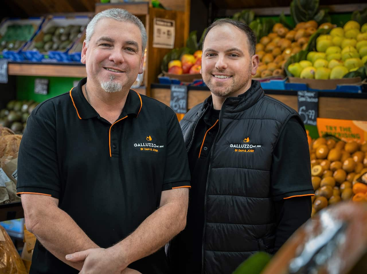 Two men in black t-shirts pose for a photo inside a fruit shop.