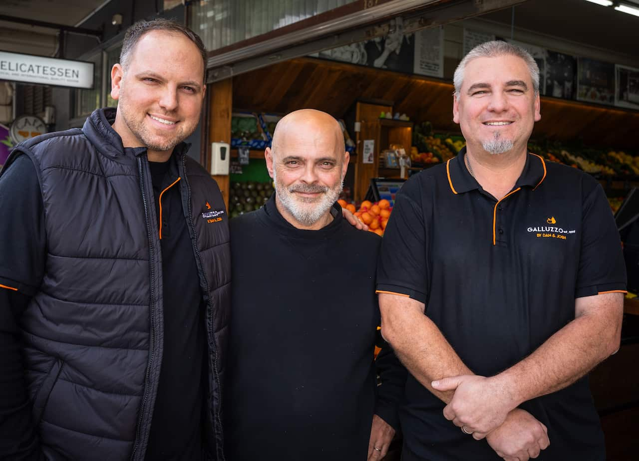 Three men in black sweaters stand together outside a fruit shop.