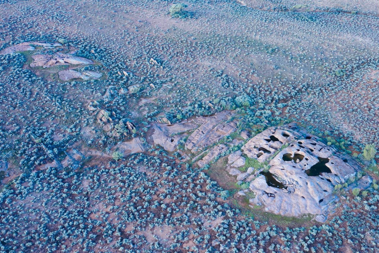 A rock hole surrounded by red dirt and scrub is seen from the air.