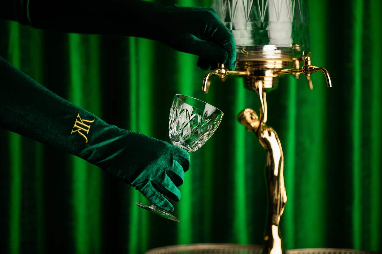 A person wearing green gloves pours a drink into a crystal goblet in front of a green backdrop