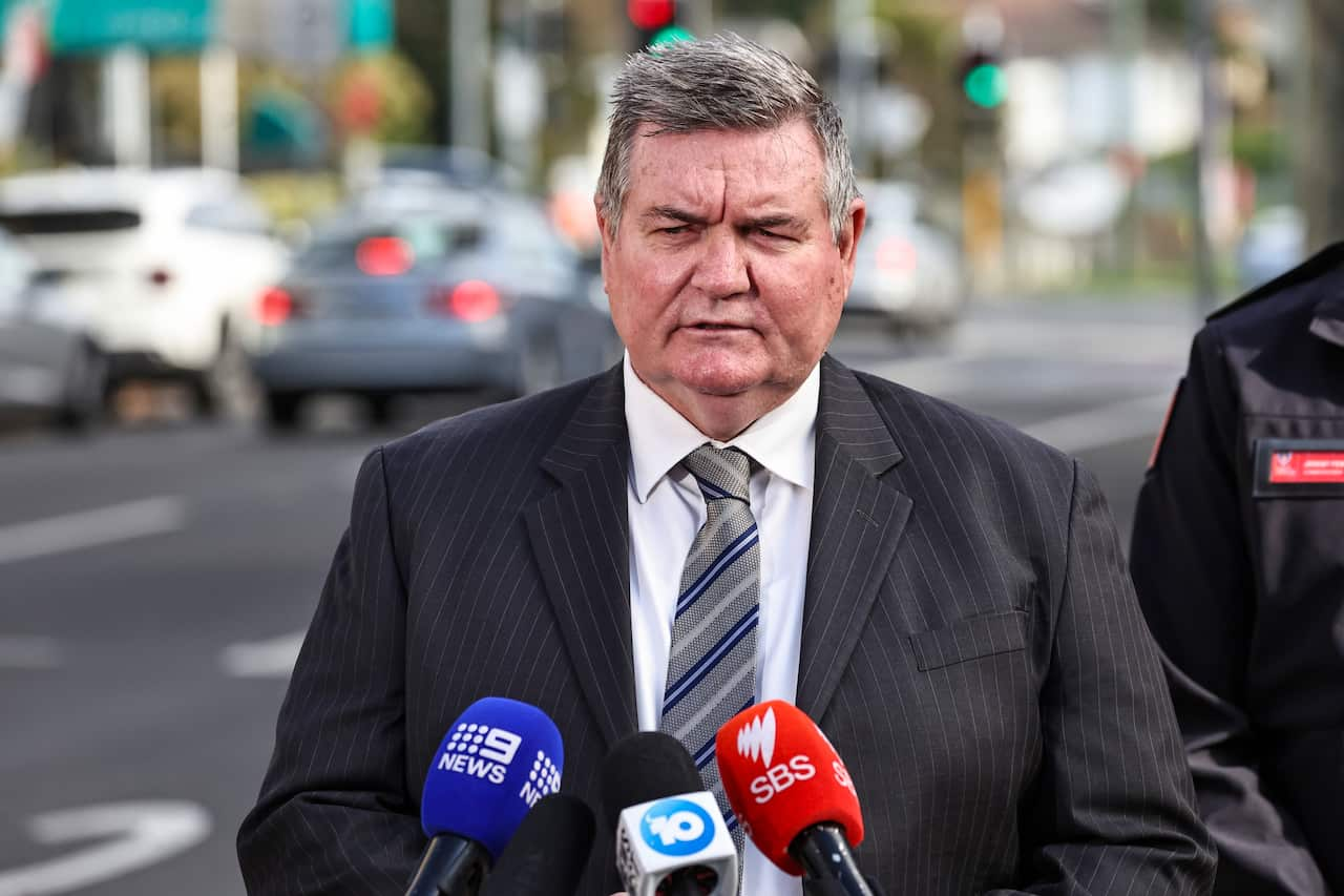 A man speaking in front of media microphones, with cars on the road behind him.