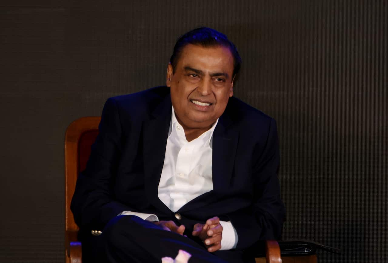 A man in a dark blazer and white shirt sitting on a brown wooden chair.