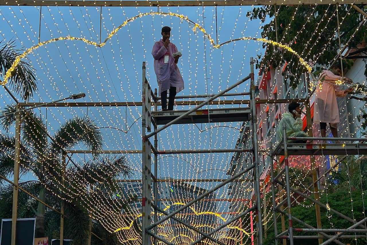 Workers standing on metal platforms decorate a venue, there are strings of lights behind them.