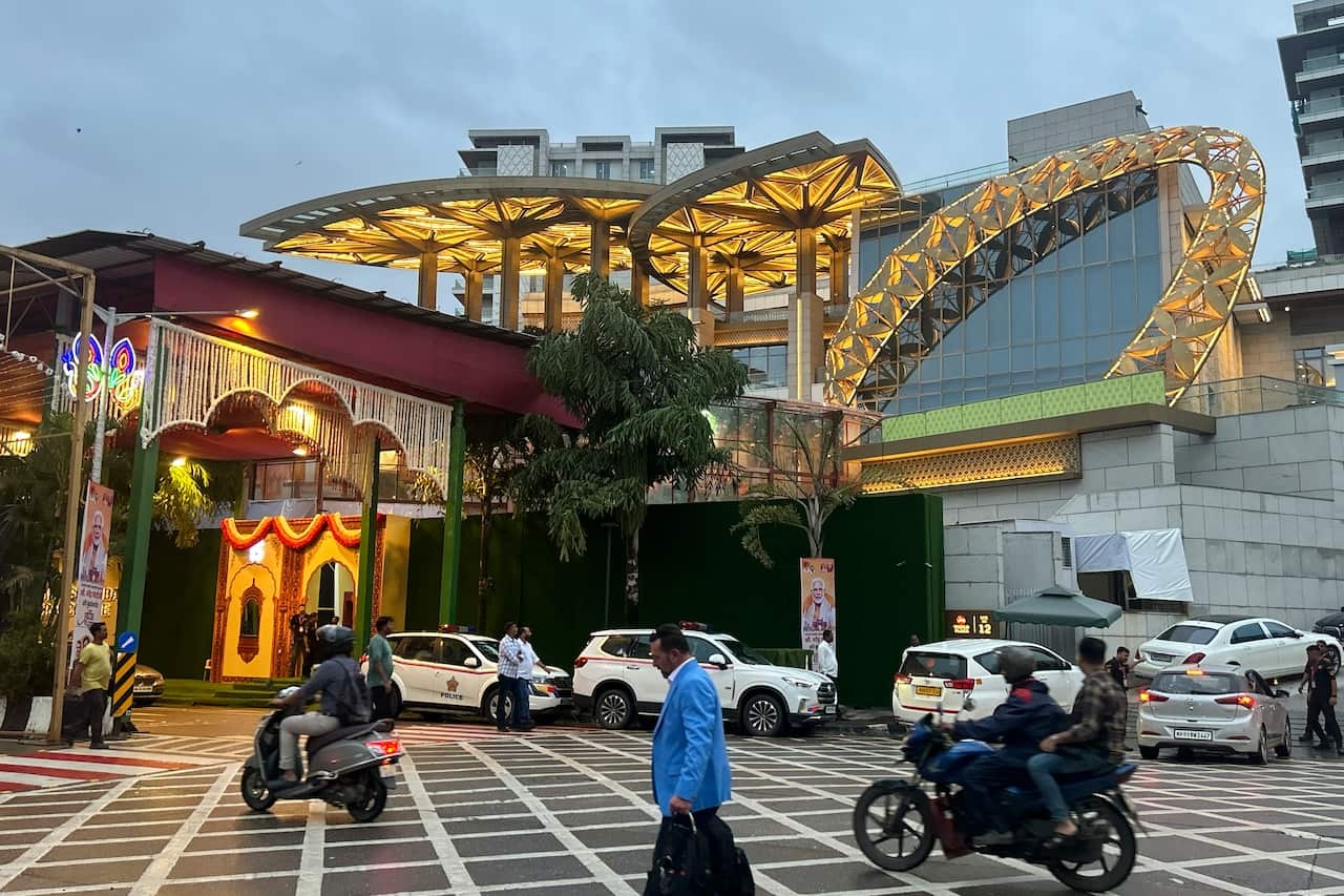 A group of people, some on motorbikes, going past an elaborate wedding venue. There are parked cars in the background.