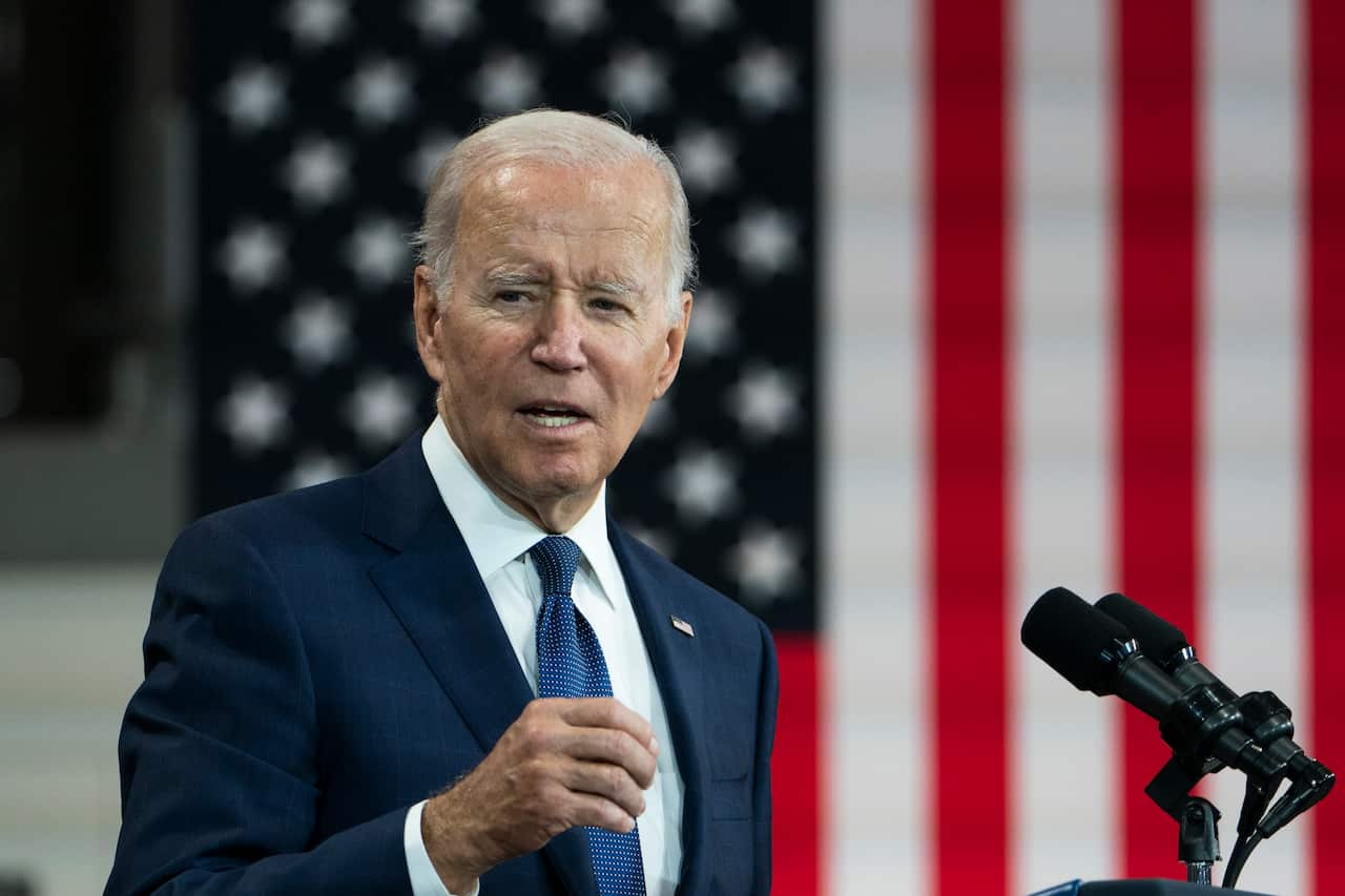 Joe Biden speaking in front of a microphone. A large American flag is behind him.