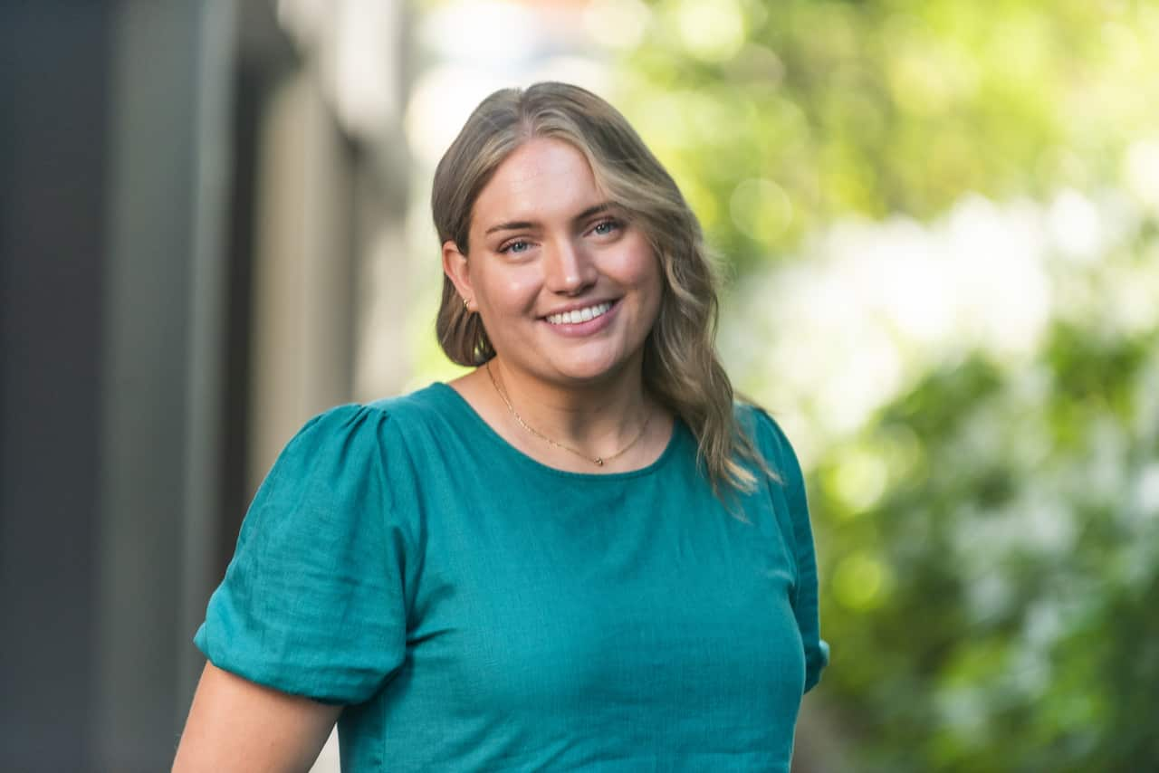 A blonde woman in a teal shirt in front of a blurred green tree. 