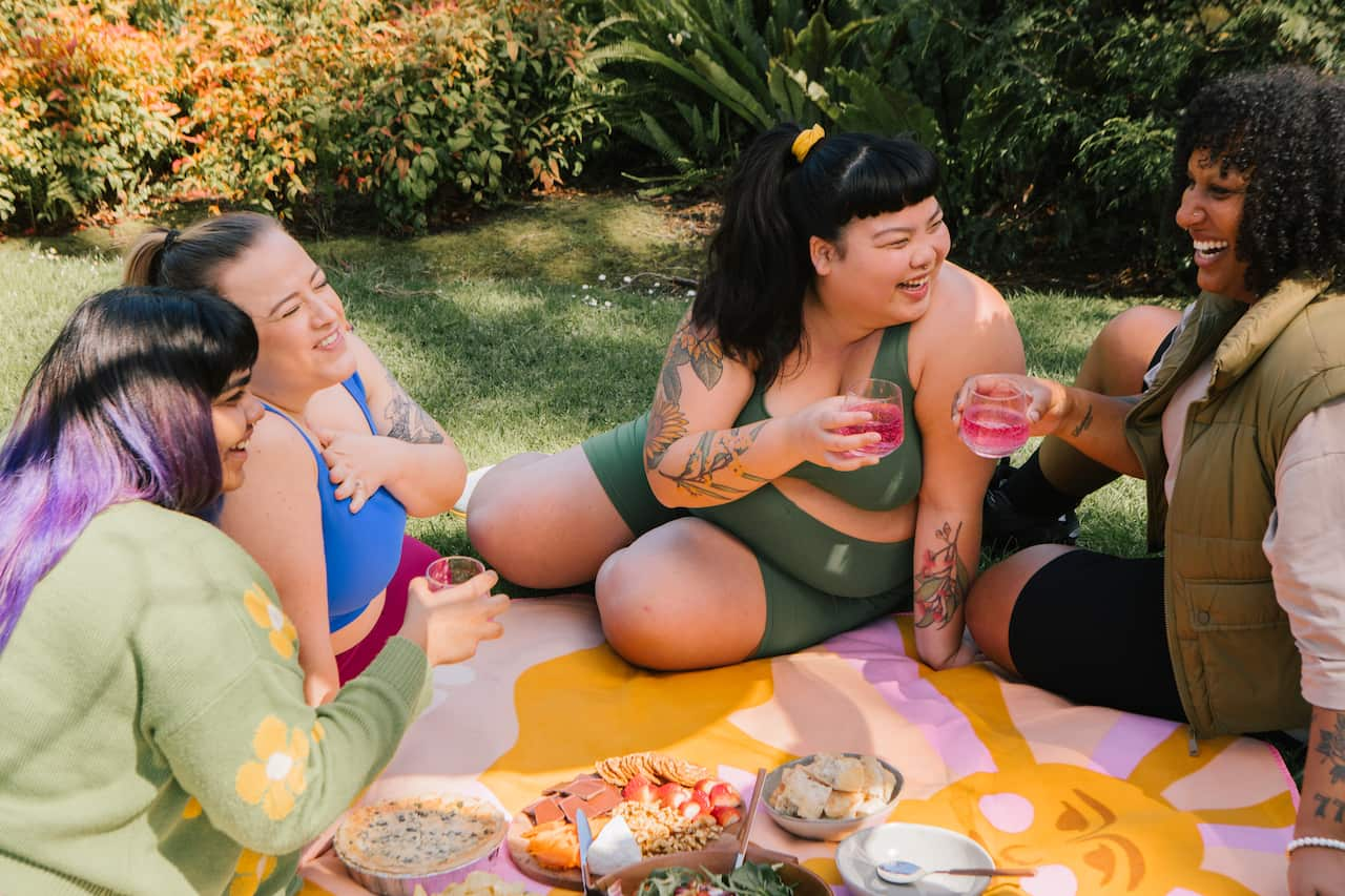 Four smiling women wearing activewear are holding drinks while sitting on a picnic blanket.