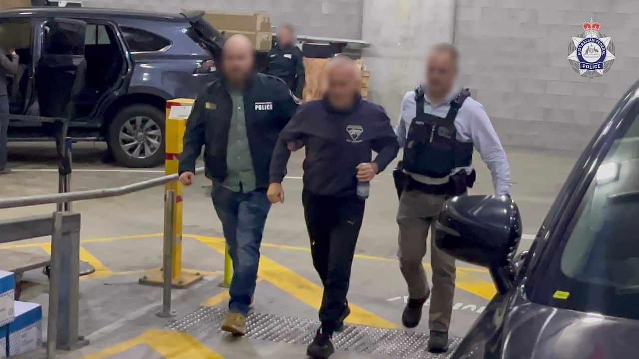 Two police officers lead a man through an underground car park. All their faces are blurred. 