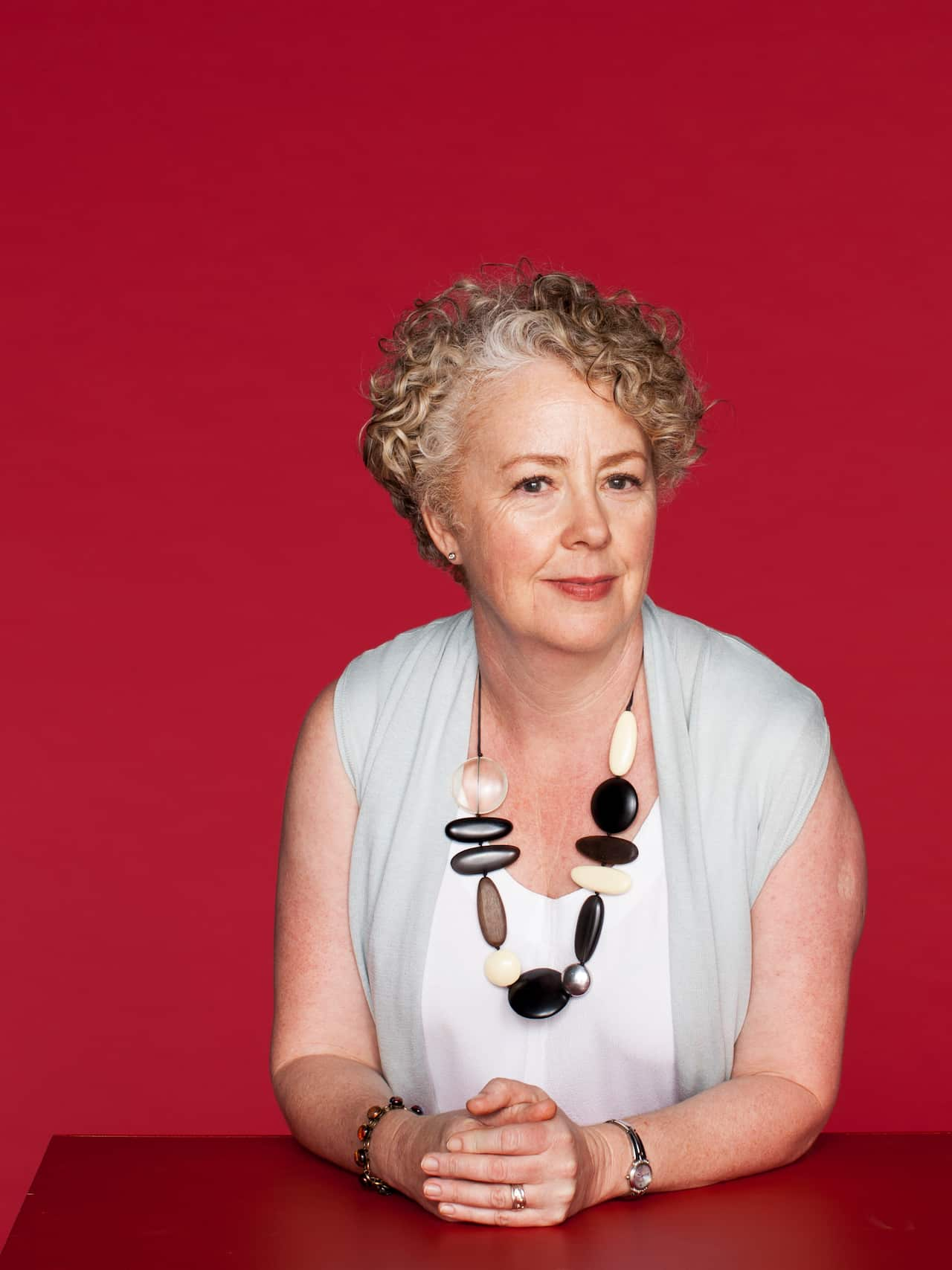 A woman with curly hair wearing a white top posing for a photograph against a crimson background