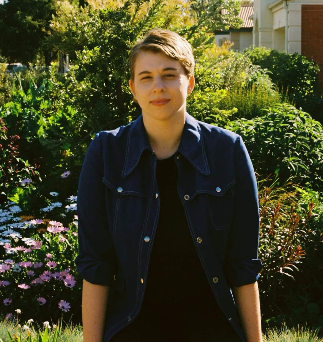 A woman with short hair wearing a denim collared shirt poses for a photograph in front of a garden. 