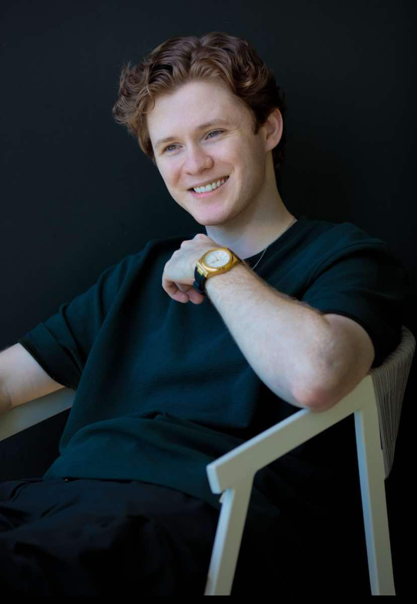 A young man wearing a dark green T-shirt sits back in a white, plastic chair. 