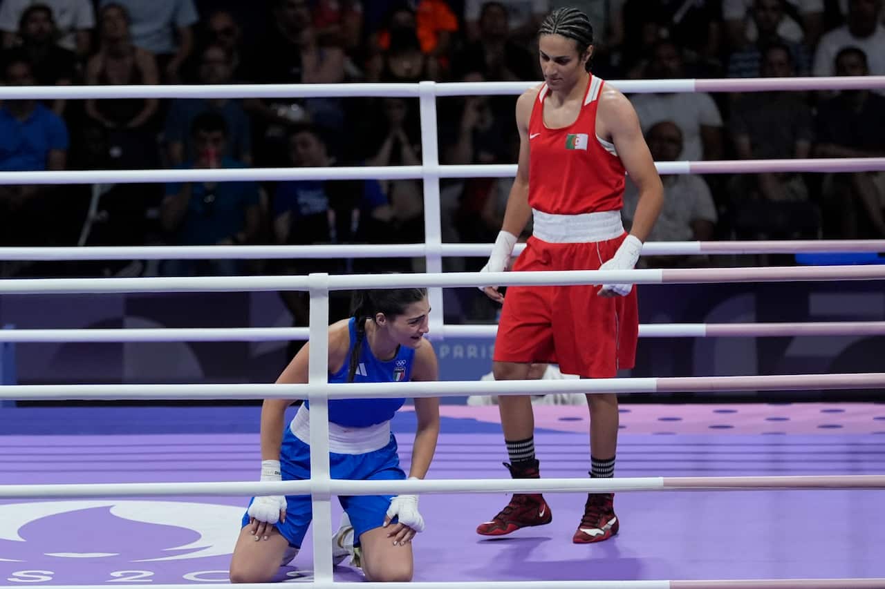 A photo of two female boxers in a boxing ring. One is kneeling on the ground and the other is standing over her. 