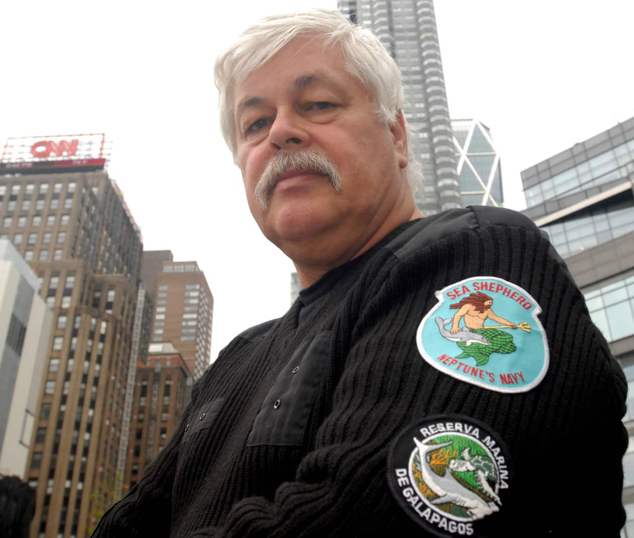 A man with grey hair and a green mustache in a black knitted jumper with a Sea Shepherd patch on his left arm.