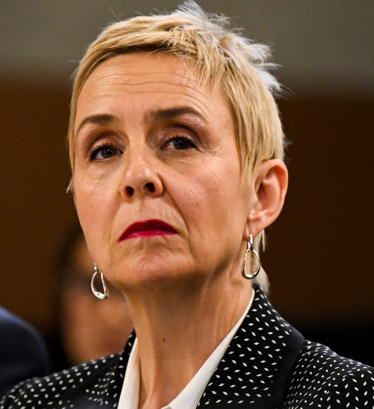 A woman with short blonde hair and a black and white dotted jacket looks to her left with a serious expression on her face. 