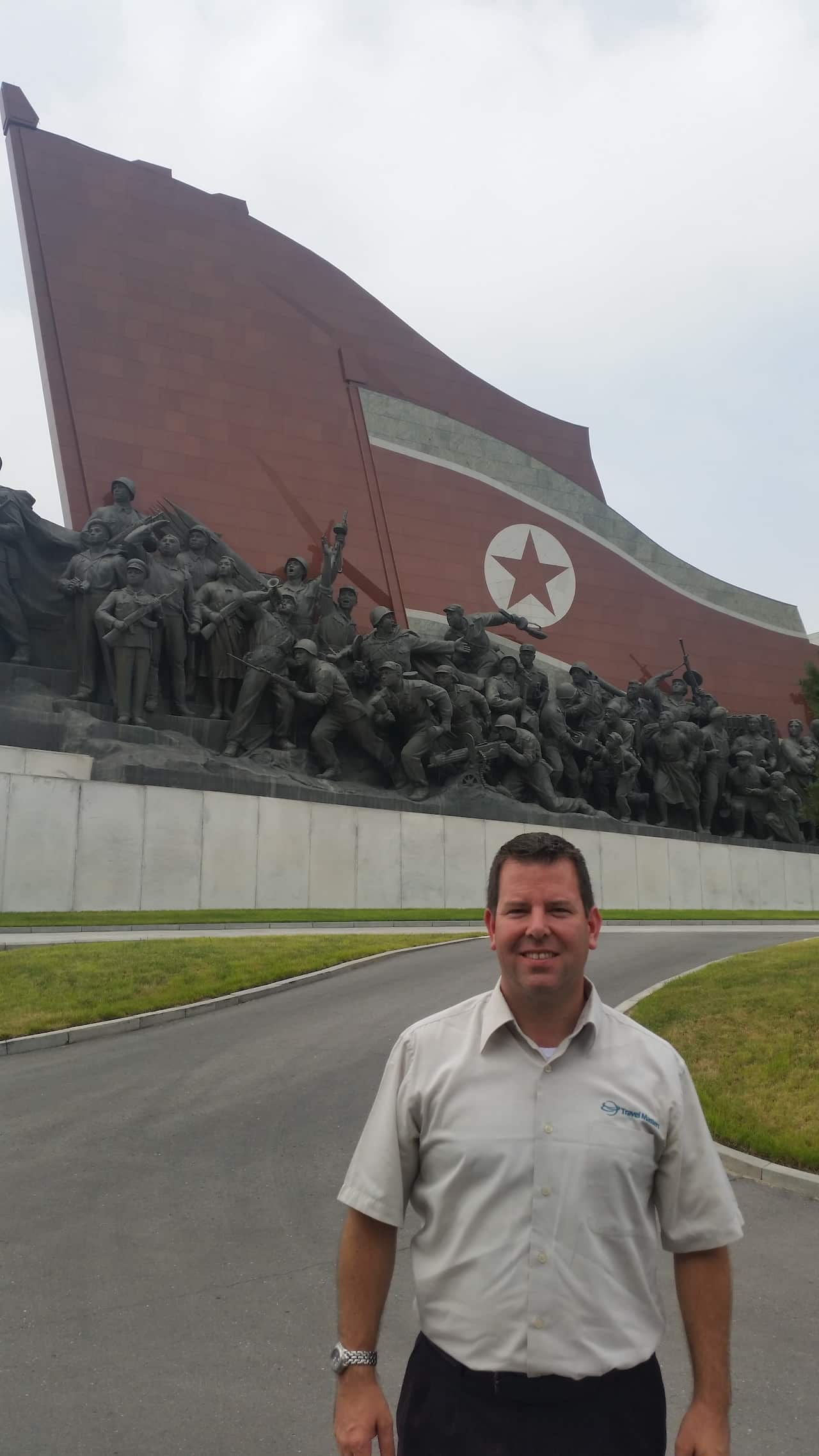 A man stands in front of a war memorial and smiles.