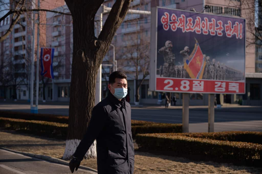 A man wearing a mask walks along a street in Pyongyang in front of a military poster.