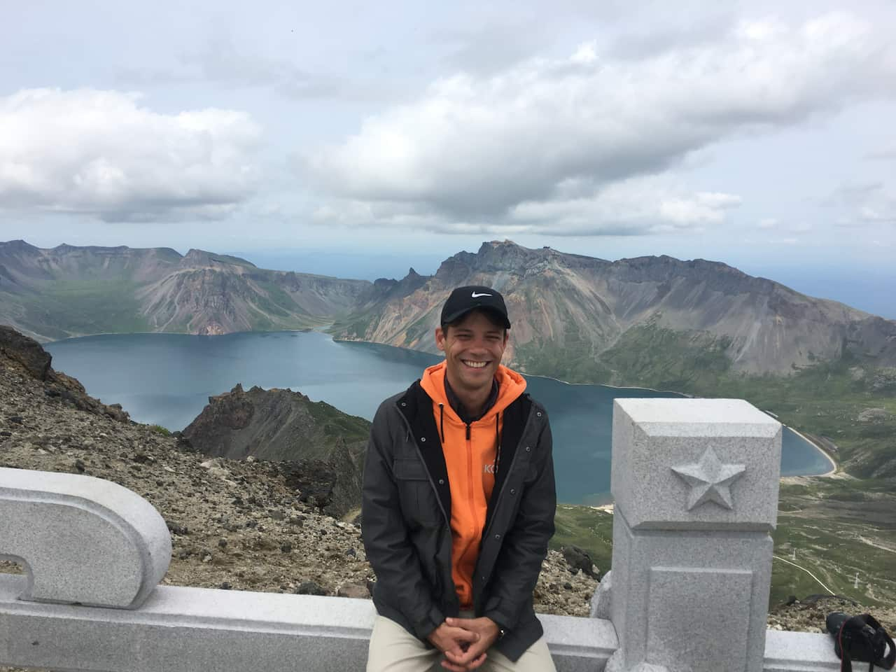 A man sits on a wall and smiles with a North Korean lake and mountains behind him.