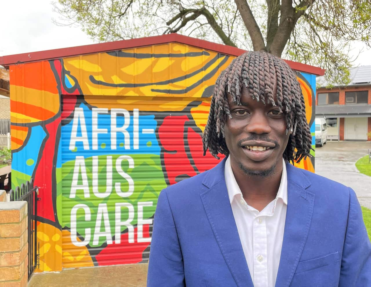 A man in a blue jacket stands in front of a garage with a colourful mural.