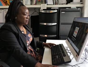 A woman in a black jackets sits in front of a computer screen in an office. 