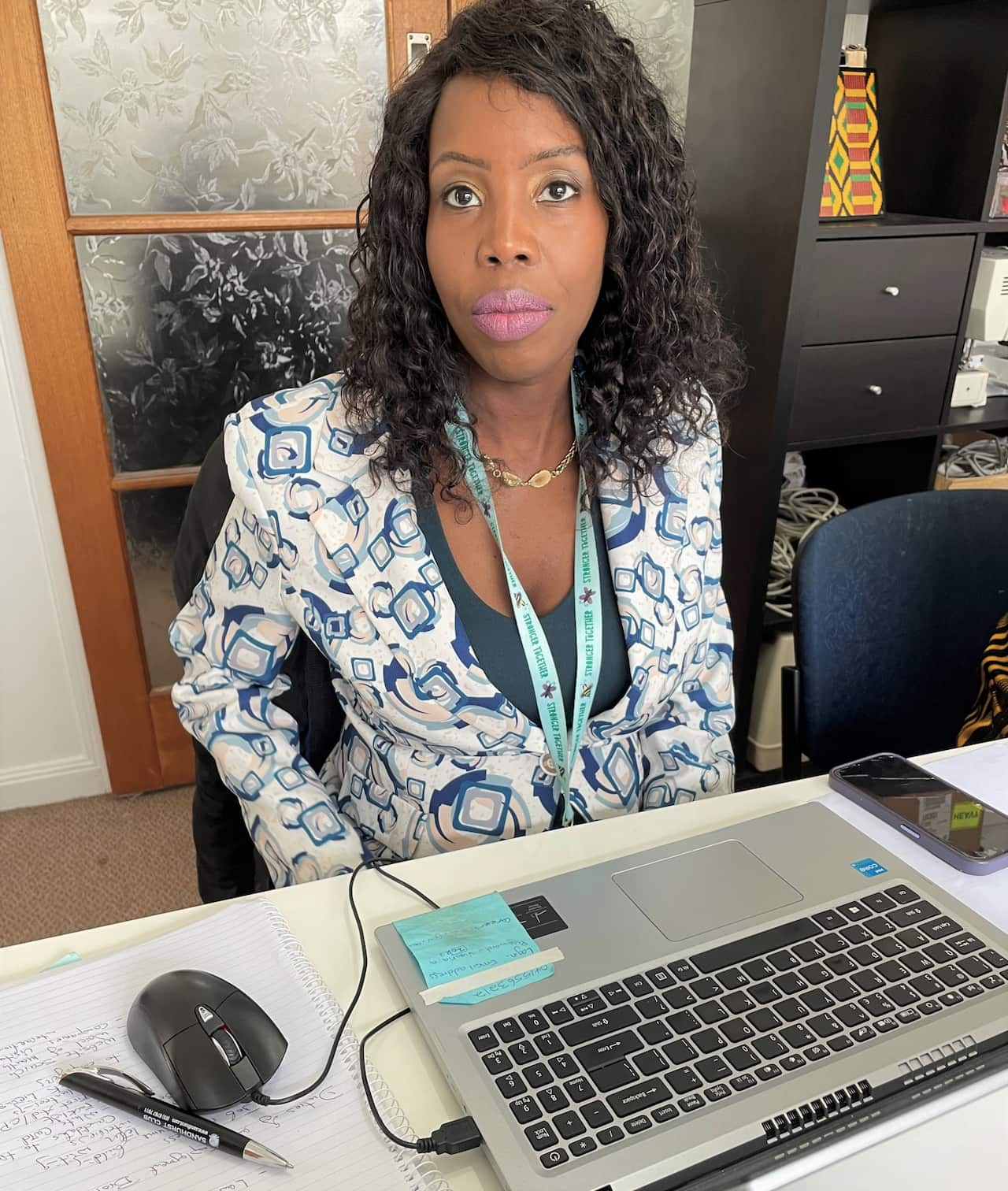 A woman in a blue and whtie jacket sits at a desk in front of a laptop.