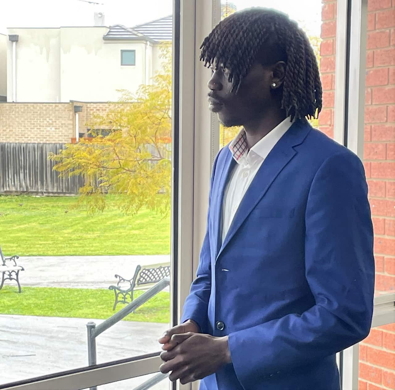 A man in a blue jacket stands at a glass window staring into a garden.